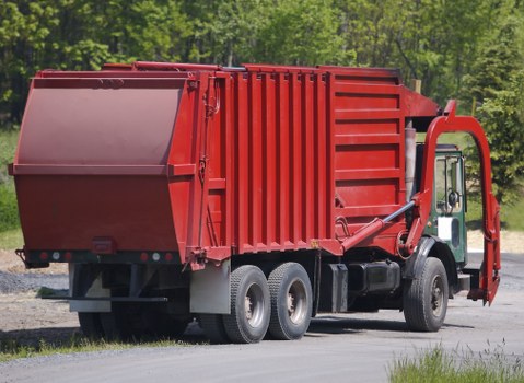 A large red waste collection truck with a rectangular, vertically ribbed metal cargo container parked on a gravel surface, with the rear opening visible and the hydraulic lifting mechanism on the side. The truck has a white cab with black wheels and tires, and is situated outdoors near a grove of green trees, suggesting a rural or suburban environment. The scene is lit with natural daylight, emphasizing the clean, metallic finish of the vehicle and the bright red colour of the container, aligning with rubbish removal services provided by Commercial Waste Golders Green. The background environment includes a grassy verge adjacent to the gravel, and the overall scene reflects a typical waste collection operation in the local area.