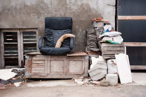 A black office chair with worn fabric upholstery and a tan cushion resting on its seat is situated on a weathered wooden chest of drawers in an outdoor, urban environment. To the right of the chair, there is a pile of construction debris, including grey concrete blocks, bricks, and white panels, stacked against a wall made of rough concrete with visible stains and marks. The background features a dark metal door and a wooden slatted panel, indicating a back alley or storage area in a commercial or residential setting. The ground is paved with concrete, scattered with smaller fragments of debris and dirt, characteristic of rubbish removal contexts in the Golders Green area. The scene appears to be part of a collection of waste, typical of rubbish collection services working in London neighborhoods, managed by Commercial Waste Golders Green. Overall, the image represents a mix of discarded furniture and construction waste, highlighting the type of material handled by professional rubbish removal services in the locality.