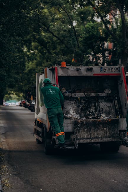 A waste collection worker wearing a green uniform and safety helmet is standing next to the open rear compartment of a large residential rubbish truck, which is parked on a quiet urban street bordered